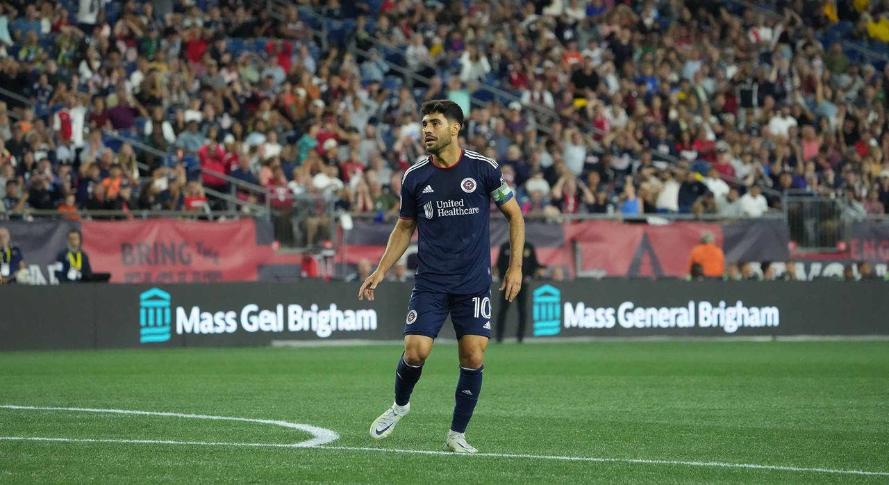 A soccer player standing by the penalty box on the pitch during a game with rows of spectators in the stadium